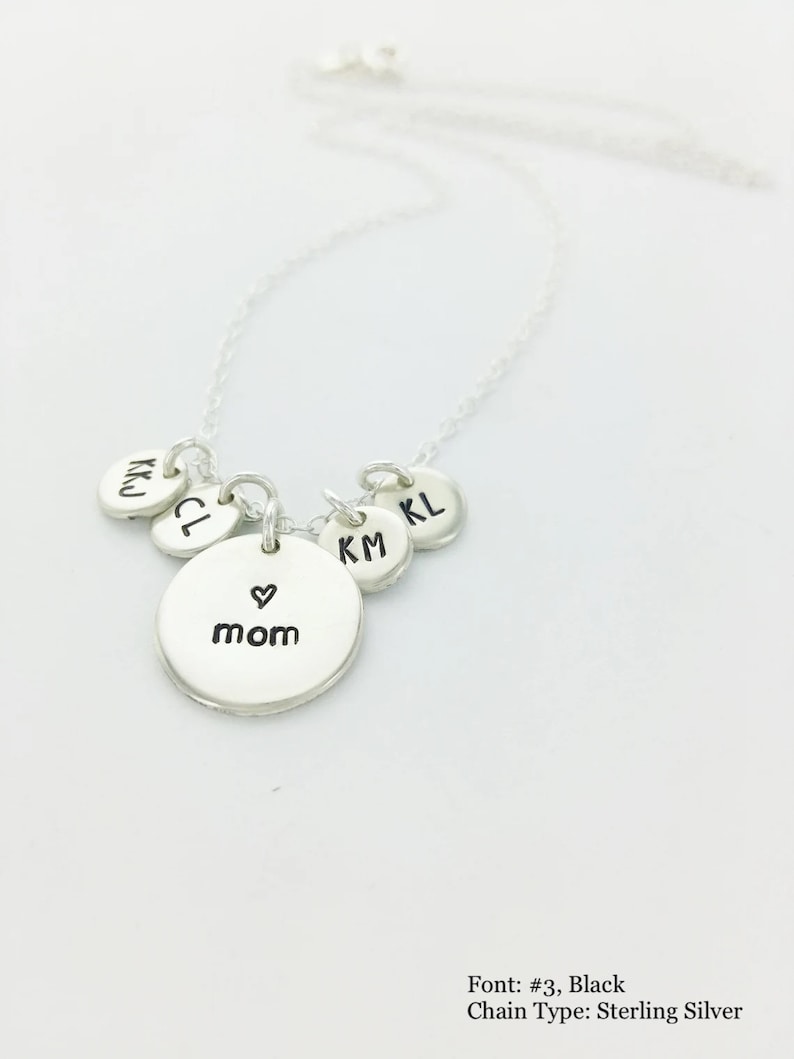 May include: A silver necklace with a delicate chain. The necklace features a central round pendant with the word "mom" and a heart symbol, along with smaller round pendants with initials. The necklace is displayed against a white background.