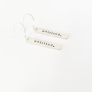 May include: A pair of silver dangle earrings with a rectangular bar pendant. Each bar has the word "untitled." engraved on it.