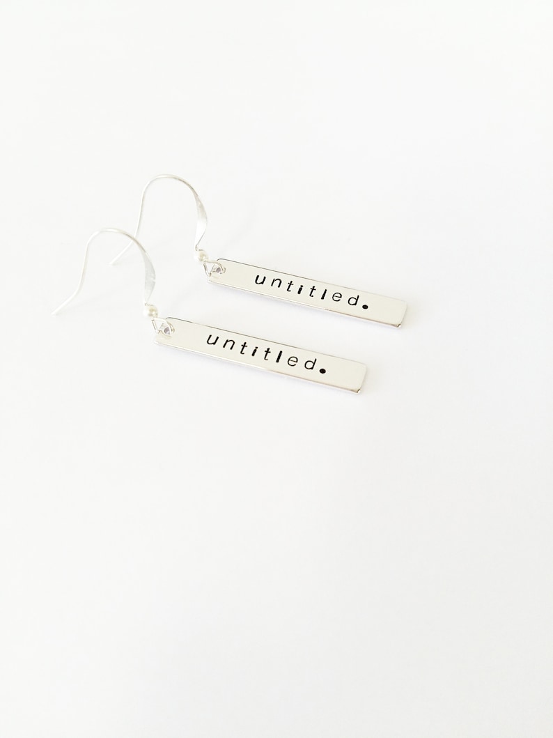 May include: A pair of silver dangle earrings with a rectangular bar pendant. Each bar has the word "untitled." engraved on it.