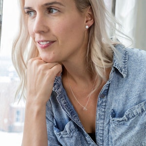 May include: A woman with blonde hair wearing a light blue denim shirt and a silver necklace with a small pendant. She is looking to the side and has her hand on her chin.