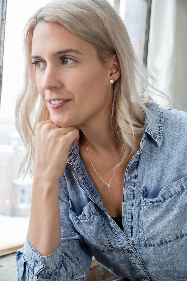 May include: A woman with blonde hair wearing a light blue denim shirt and a silver necklace with a small pendant. She is looking to the side and has her hand on her chin.