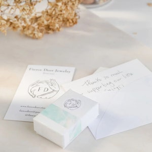 May include: A white box with a watercolor design and the letters "FD" in a circle. The box is sitting on a white surface next to a white card with the text "Fierce Deer Jewelry" and "Thanks so much supporting our little mimi!"