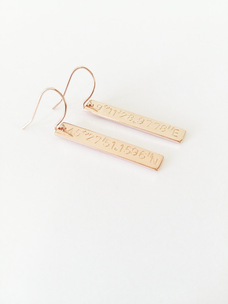May include: Rose gold earrings with a bar design. Each bar has coordinates engraved on it: 45.27751.1596&deg;N and 9&deg;11'28.9778&deg;E.