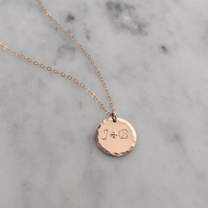 May include: Rose gold necklace with a hammered-texture pendant. The pendant is a small, round disc with the engraved initials "J+B". The chain and pendant are a warm, metallic color.