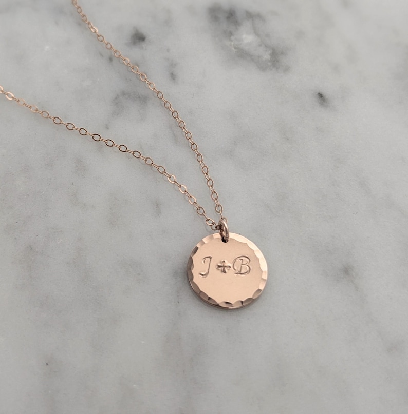 May include: Rose gold necklace with a hammered-texture pendant. The pendant is a small, round disc with the engraved initials "J+B". The chain and pendant are a warm, metallic color.