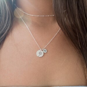 May include: A silver necklace with two pendants. One pendant is a small, round charm with a dandelion design. The other is a smaller, round charm with a single initial. A delicate chain necklace is also visible.