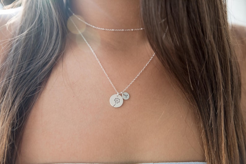 May include: A silver necklace with two pendants. One pendant is a small, round charm with a dandelion design. The other is a smaller, round charm with a single initial. A delicate chain necklace is also visible.