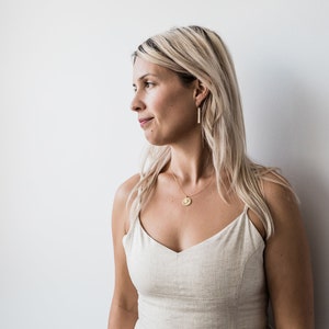 May include: A woman with blonde hair wearing a light beige sleeveless top with a gold necklace and gold earrings. The necklace has a round pendant.