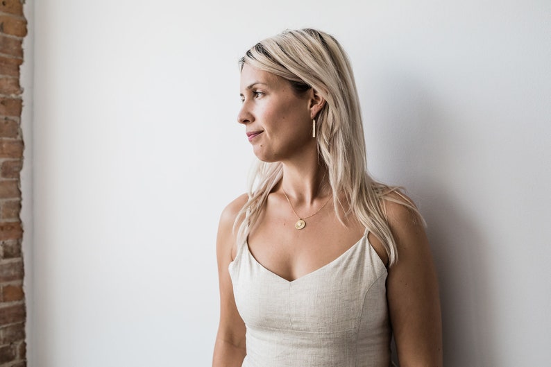May include: A woman with blonde hair wearing a light beige sleeveless top with a gold necklace and gold earrings. The necklace has a round pendant.