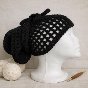 May include: A black crocheted hat with a net-like design is displayed on a white mannequin head. The hat has a knotted accent and a long, draped section. Two balls of yarn and a crochet hook are in the background.