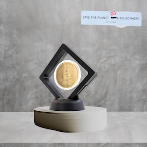 Puede incluir: Una moneda de oro exhibida en un marco flotante negro y transparente, colocada sobre un pedestal blanco y gris. La moneda está centrada dentro del marco. Una pegatina en la parte superior dice "SAVE THE PLANET, BE A BILLIONAIRE."