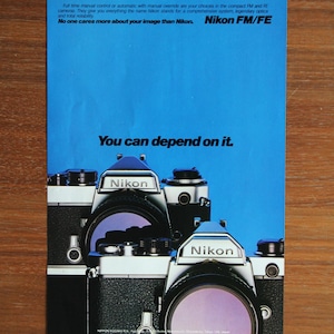 Peut inclure: Publicité vintage pour les appareils photo Nikon FM/FE. L'annonce présente trois appareils photo sur fond bleu avec le logo Nikon. Le texte comprend "You can depend on it."