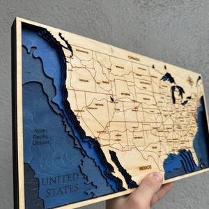 May include: A multi-layered wooden map of the United States featuring a light wood finish for landmasses and deep blue, tiered cutouts representing the Pacific Ocean depths. The map includes engraved state names, borders, and the North Pacific Ocean label.