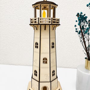 May include: A laser-cut wooden lighthouse model featuring a hexagonal base, multiple levels with arched windows, and a top gallery railing. The structure is light tan with dark brown edges and contains a small, glowing yellow LED light inside.