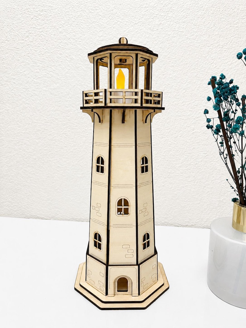 May include: A laser-cut wooden lighthouse model featuring a hexagonal base, multiple levels with arched windows, and a top gallery railing. The structure is light tan with dark brown edges and contains a small, glowing yellow LED light inside.