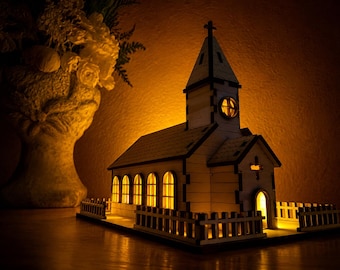 May include: A miniature wooden church model featuring a tall steeple with a cross, arched windows, and a surrounding picket fence. The structure is illuminated from within by a warm yellow light, casting a glow through the windows and doorway.