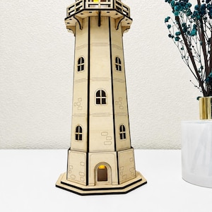 May include: A laser-cut wooden lighthouse model featuring a multi-tiered structure with dark-edged panels, arched windows, and a balcony. The tower includes a small internal yellow light visible at the top and base, with engraved rectangular brick patterns on the exterior walls.