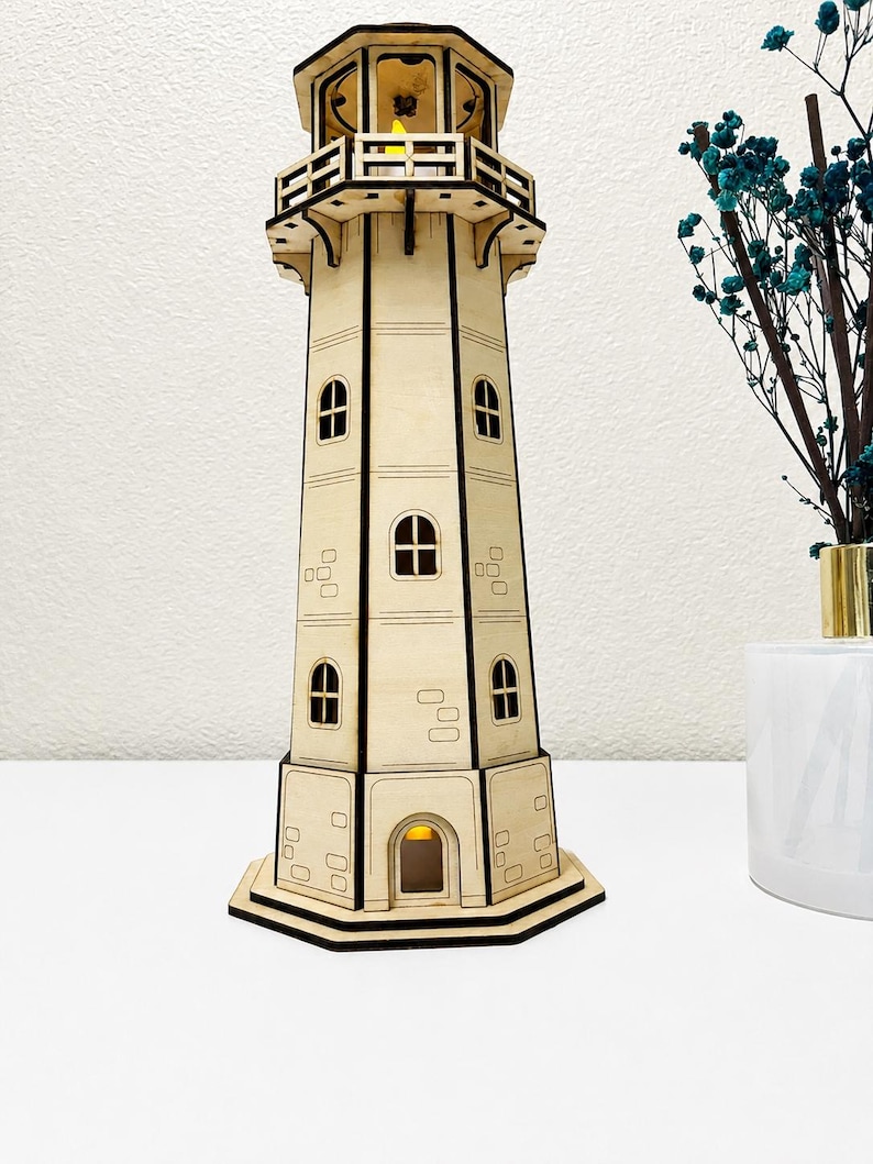 May include: A laser-cut wooden lighthouse model featuring a multi-tiered structure with dark-edged panels, arched windows, and a balcony. The tower includes a small internal yellow light visible at the top and base, with engraved rectangular brick patterns on the exterior walls.