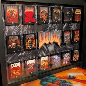 Puede incluir: Una exhibición enmarcada con varias cajas de juego DOOM en miniatura con diferentes portadas. La palabra "DOOM" se muestra prominentemente en una fuente 3D naranja y plateada. El marco es negro y el fondo tiene un patrón oscuro y texturizado.