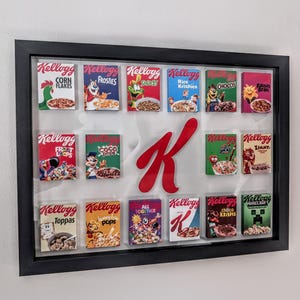 Op de afbeelding: Een ingelijste display met miniatuur Kellogg's ontbijtgranen dozen. De dozen, in verschillende kleuren, tonen verschillende ontbijtgranen zoals Frosties, Corn Flakes en Rice Krispies. Een groot rood "K" logo is gecentreerd en voegt een opvallend accent toe.