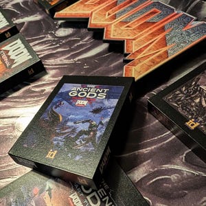 Op de afbeelding: Verschillende zwarte speldozen met het "DOOM" logo en de tekst "Eternal" worden getoond. E&eacute;n doos toont artwork van "The Ancient Gods Part One". De dozen hebben het "id" logo. De achtergrond is een patroon.