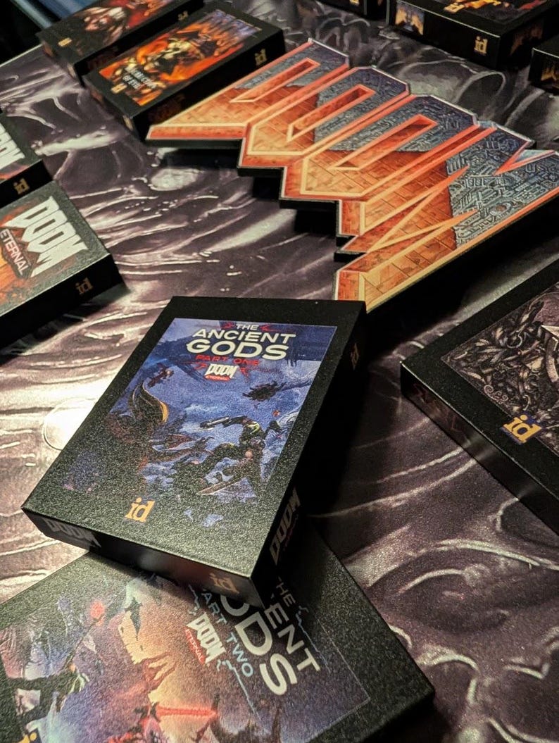Op de afbeelding: Verschillende zwarte speldozen met het "DOOM" logo en de tekst "Eternal" worden getoond. E&eacute;n doos toont artwork van "The Ancient Gods Part One". De dozen hebben het "id" logo. De achtergrond is een patroon.
