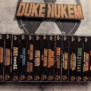 May include: A collection of Duke Nukem video game boxes, featuring various titles like "Duke Nukem 3D" and "Duke Nukem: Manhattan Project." The boxes are black with gold lettering and graphics, and the "Duke Nukem" logo is displayed above.