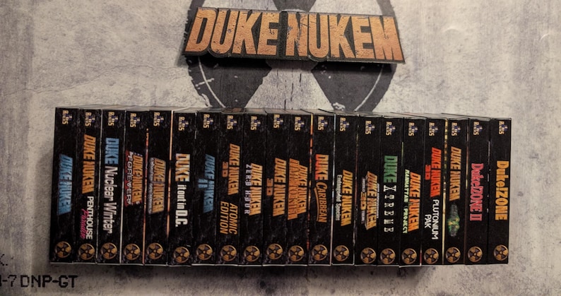 May include: A collection of Duke Nukem video game boxes, featuring various titles like "Duke Nukem 3D" and "Duke Nukem: Manhattan Project." The boxes are black with gold lettering and graphics, and the "Duke Nukem" logo is displayed above.