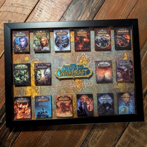 Puede incluir: Un expositor enmarcado en negro con cajas de juego World of Warcraft en miniatura. Las cajas están dispuestas sobre un fondo de mapa con el logotipo del juego. El conjunto es un objeto de colección.