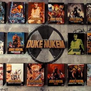 May include: A collection of Duke Nukem video game boxes, featuring various action-packed cover art. The boxes display the game's logo and character in dynamic poses, with a variety of color schemes and graphic designs. The collection is arranged on a textured background.