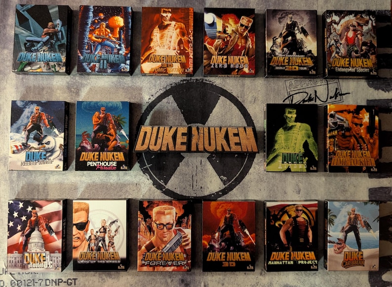 May include: A collection of Duke Nukem video game boxes, featuring various action-packed cover art. The boxes display the game's logo and character in dynamic poses, with a variety of color schemes and graphic designs. The collection is arranged on a textured background.