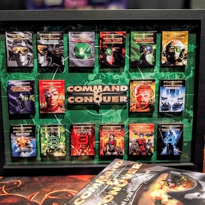 May include: A framed collection of Command & Conquer game boxes. The boxes are arranged on a green background, with the game title prominently displayed. The boxes feature various character portraits and game titles.