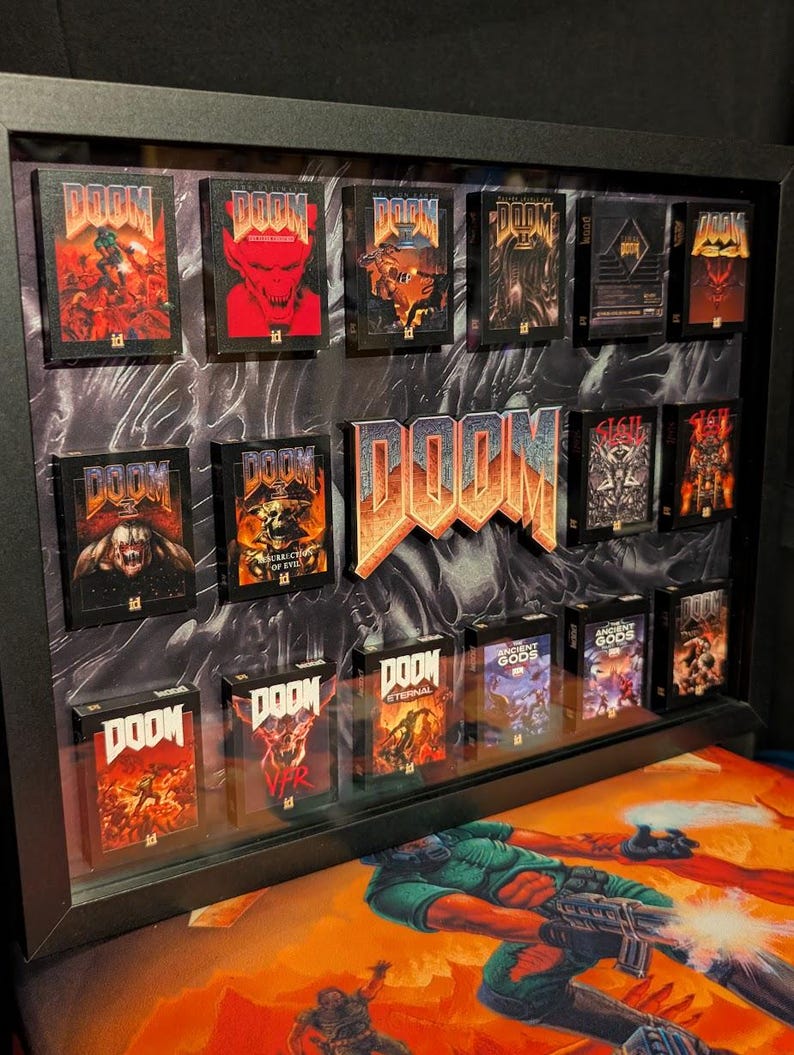 Op de afbeelding: Een ingelijste display met miniatuur DOOM-gameboxen met diverse cover art. Het woord "DOOM" is prominent weergegeven in een 3D-lettertype in oranje en zilver. De lijst is zwart en de achtergrond heeft een donker, getextureerd patroon.