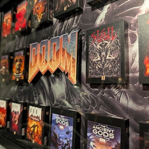 Op de afbeelding: Een ingelijste display met miniatuur DOOM-gameboxen en een 3D DOOM-logo. De dozen tonen verschillende speltitels met levendige illustraties. Het logo is oranje en blauw en de achtergrond heeft een getextureerd, donker ontwerp.