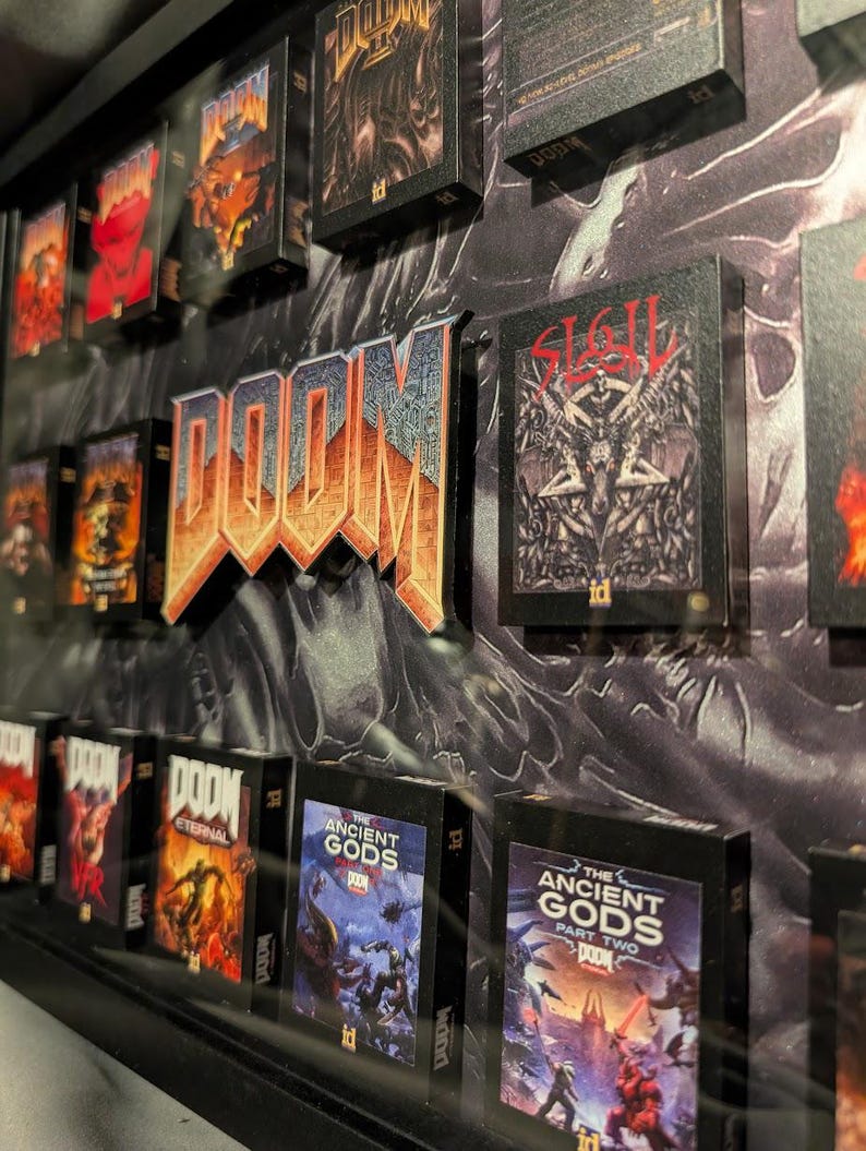 Op de afbeelding: Een ingelijste display met miniatuur DOOM-gameboxen en een 3D DOOM-logo. De dozen tonen verschillende speltitels met levendige illustraties. Het logo is oranje en blauw en de achtergrond heeft een getextureerd, donker ontwerp.