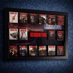 Puede incluir: Una exhibición enmarcada en negro con múltiples portadas de juegos de Resident Evil. El marco contiene varias cajas de juegos con diferentes ilustraciones, incluyendo imágenes de personajes, zombis y el logotipo del juego. El fondo muestra un paisaje urbano y las palabras "Resident Evil" y "Raccoon City".