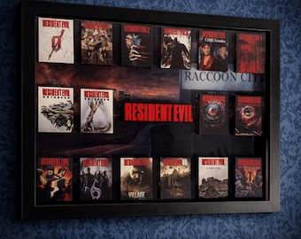 May include: A framed display featuring multiple Resident Evil game covers. The black frame holds various game boxes with different cover art, including images of characters, zombies, and the game's logo. The background shows a cityscape and the words "Resident Evil" and "Raccoon City".