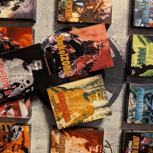 May include: A collection of Duke Nukem comic books and video game boxes. The boxes feature action-oriented illustrations in various colors, including red, green, and yellow. The text "Duke Nukem" and "Duke!Zone" are visible on the boxes.