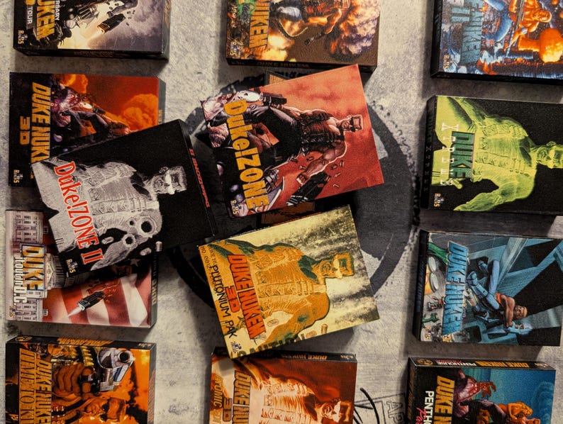 May include: A collection of Duke Nukem comic books and video game boxes. The boxes feature action-oriented illustrations in various colors, including red, green, and yellow. The text "Duke Nukem" and "Duke!Zone" are visible on the boxes.