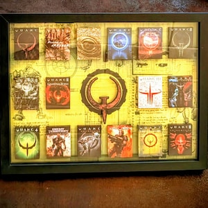 Peut inclure: Collection encadrée de pochettes de jeux vidéo Quake. Le cadre noir entoure un fond jauni avec diverses pochettes de jeux, présentant le logo Quake et les titres des jeux. Les pochettes affichent une gamme de couleurs et de graphismes.