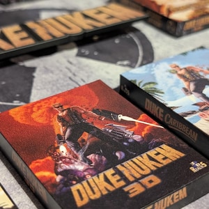 May include: Multiple game boxes featuring the "Duke Nukem" series. The boxes have a dark background with the game title in orange and images of the game characters. The "Duke Nukem 3D" box has a red and orange background.