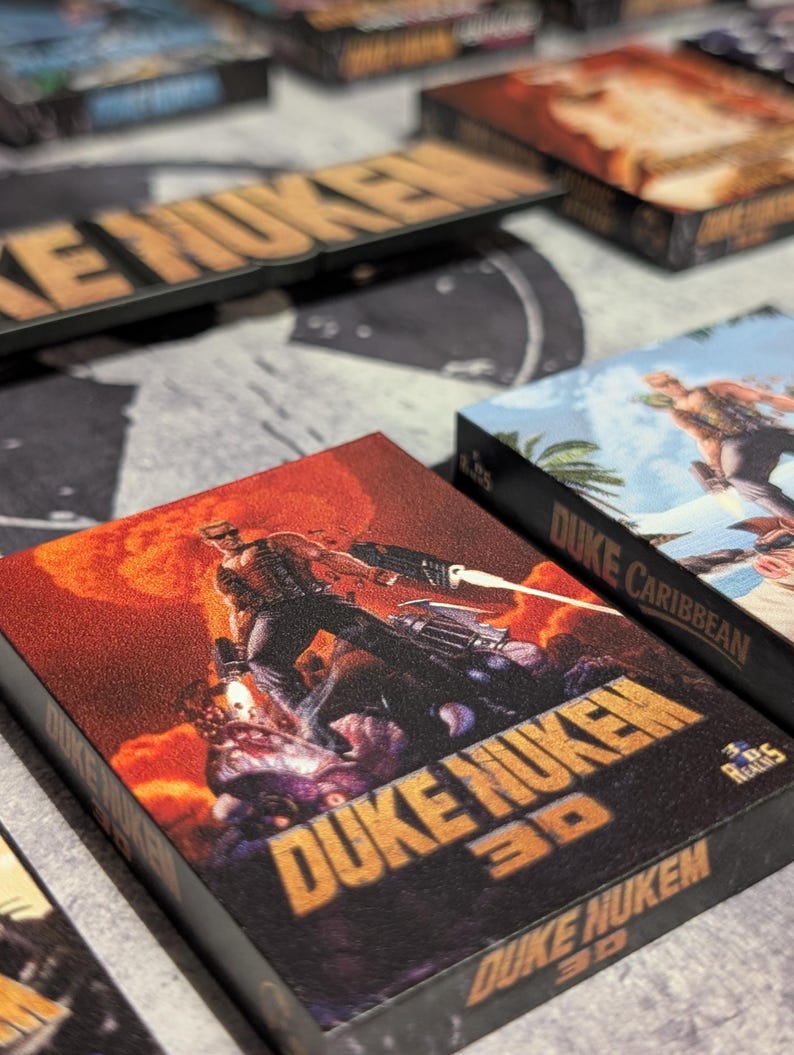May include: Multiple game boxes featuring the "Duke Nukem" series. The boxes have a dark background with the game title in orange and images of the game characters. The "Duke Nukem 3D" box has a red and orange background.