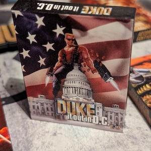 May include: A game box featuring a muscular character with sunglasses, holding weapons, and standing in front of the U.S. Capitol building. The background is an American flag. The text "DUKE it out in D.C." is displayed.