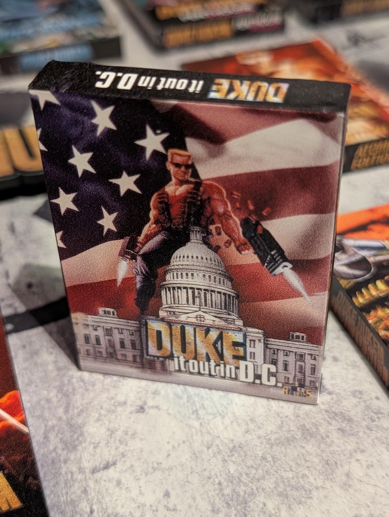 May include: A game box featuring a muscular character with sunglasses, holding weapons, and standing in front of the U.S. Capitol building. The background is an American flag. The text "DUKE it out in D.C." is displayed.