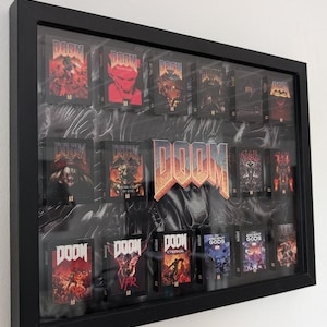 Op de afbeelding: Een zwart omkaderde display met meerdere DOOM game cartridges. De cartridges hebben diverse cover art, waaronder afbeeldingen van demonen en actiesc&egrave;nes. De achtergrond heeft een marmer effect. Het woord "DOOM" is prominent weergegeven.