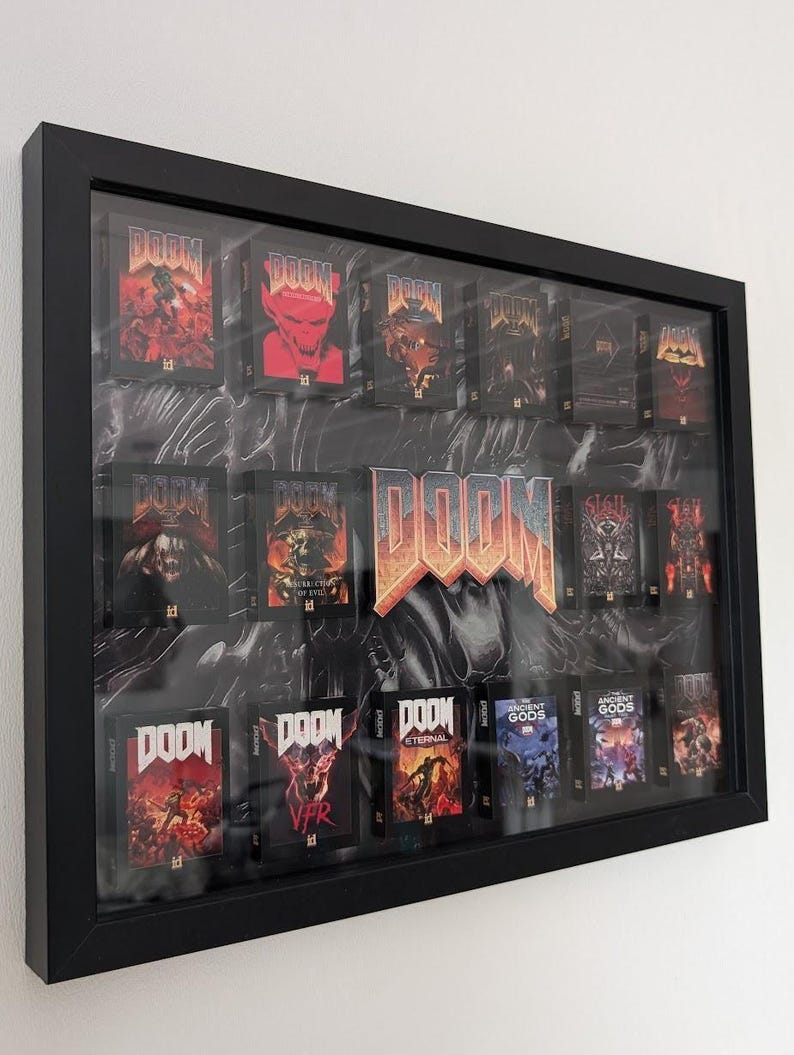Op de afbeelding: Een zwart omkaderde display met meerdere DOOM game cartridges. De cartridges hebben diverse cover art, waaronder afbeeldingen van demonen en actiesc&egrave;nes. De achtergrond heeft een marmer effect. Het woord "DOOM" is prominent weergegeven.
