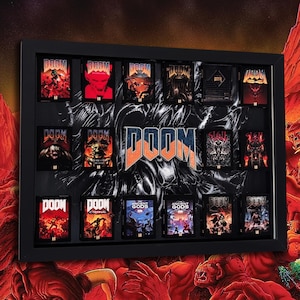 May include: A black framed display case featuring a collection of sixteen Doom video game covers arranged in a grid. The centre displays the Doom logo in blue and orange. Each individual game cover showcases distinct dark, red, and orange artwork.