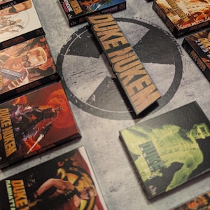 May include: A collection of Duke Nukem video game boxes and a wooden sign with the game's logo. The boxes feature various game covers with action-oriented illustrations and the game's title. The sign displays the game's name in a bold, distressed font.