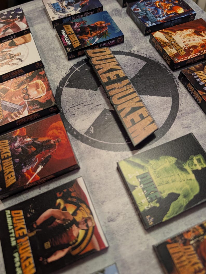 May include: A collection of Duke Nukem video game boxes and a wooden sign with the game's logo. The boxes feature various game covers with action-oriented illustrations and the game's title. The sign displays the game's name in a bold, distressed font.