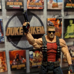 May include: A Duke Nukem action figure in a red tank top and black pants, holding a gun. The figure is in front of a Duke Nukem logo and several game box covers.